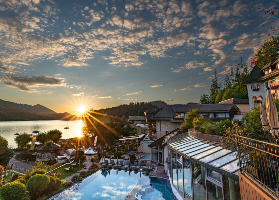 Sunset view over Waldhof Fuschlsee Resort with an infinity pool, lake, and mountains in the background.