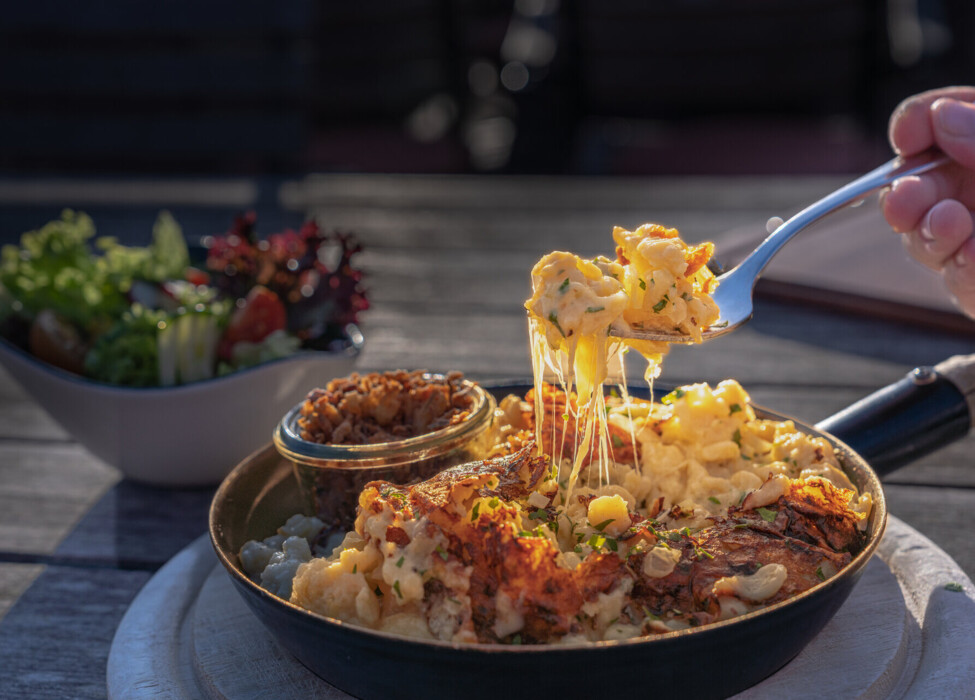 Kasnockn mit geschmolzenem Käse im Pfännchen, serviert mit einem frischen Salat im Hintergrund auf einem Tisch.