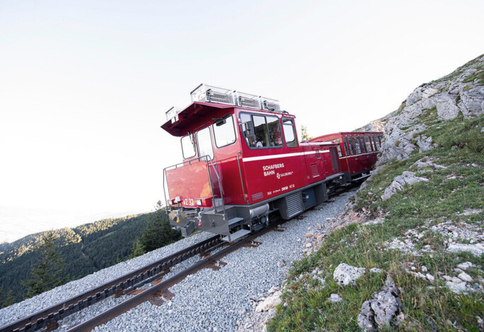 Roter Zug der Schafbergbahn fährt auf einer Bergstrecke, umgeben von felsiger Landschaft und Bäumen, unter klarem Himmel.