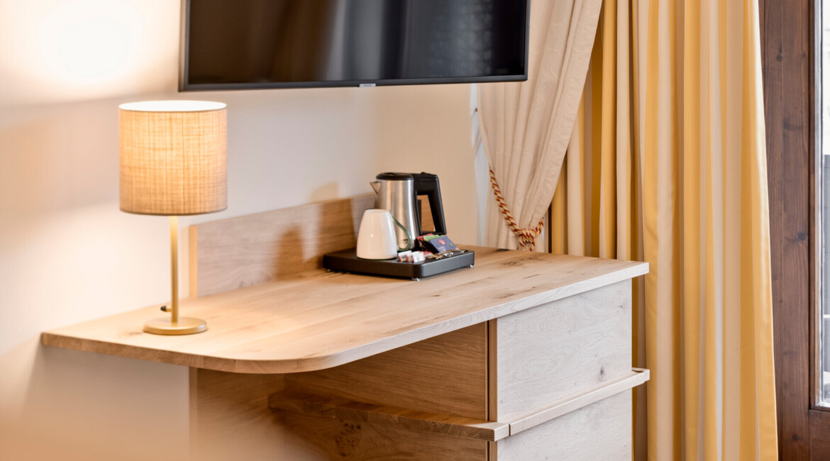 A cozy hotel room corner with a wooden desk, lamp, TV, and kettle set against cream curtains.