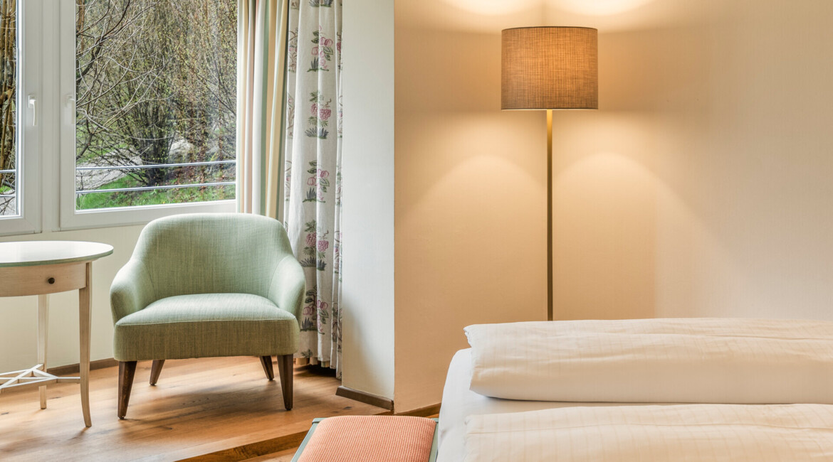 A cozy hotel room with a comfortable chair, a floor lamp, and a view of greenery through large windows.