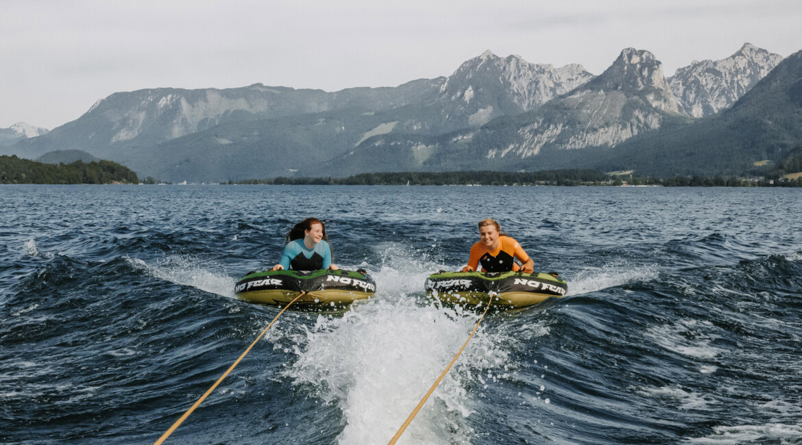 Two people enjoying tubing on a lake with mountain views in the background.