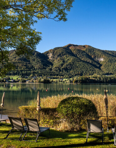 Lakeside loungers facing a mountain view at Waldhof Fuschlsee Resort, surrounded by greenery under a clear blue sky.