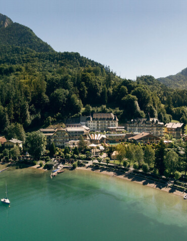 Aerial view of Waldhof Fuschlsee Resort by a tranquil lake, surrounded by forested hills and mountains.