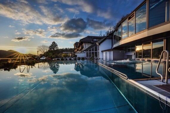 Outdoor pool reflecting the sunset and clouds, with a modern building and natural landscape in the background.