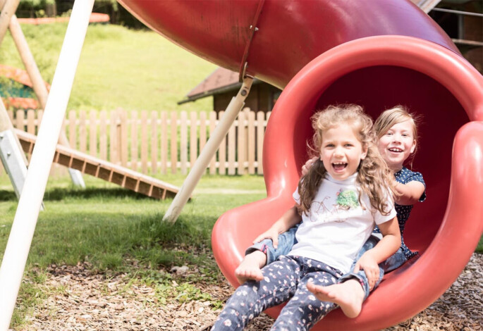 Children happily sliding down a red slide at a playground with a scenic, grassy background.