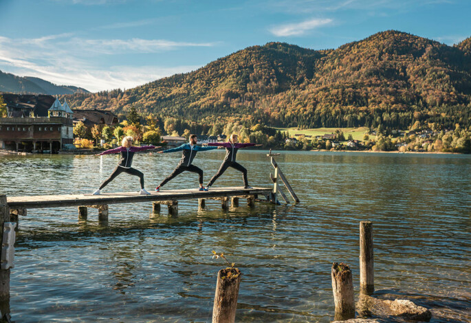 Three people practicing yoga on a dock by a picturesque lake, surrounded by lush mountains.