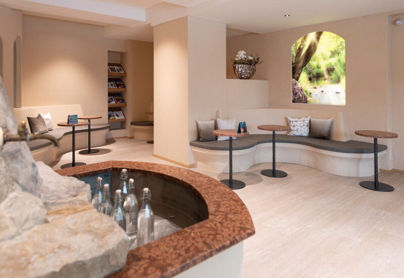 Cozy seating area with curved benches and small tables, featuring a scenic window view of lush greenery and a stone fountain with water bottles.