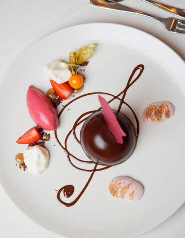 Elegantes Dessert mit Schokoladenkuppel, Beeren und Sahne, kunstvoll auf einem weißen Teller angerichtet.