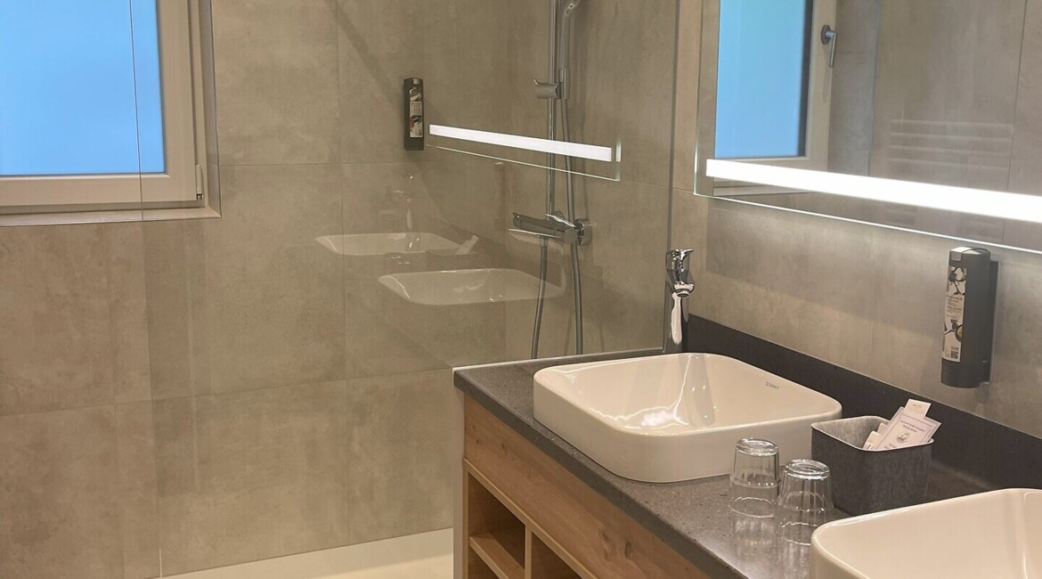 Modernes Badezimmer mit großer ebenerdiger Dusche, zwei Waschbecken und Spiegel. Elegantes Design mit Holz- und Glasakzenten.