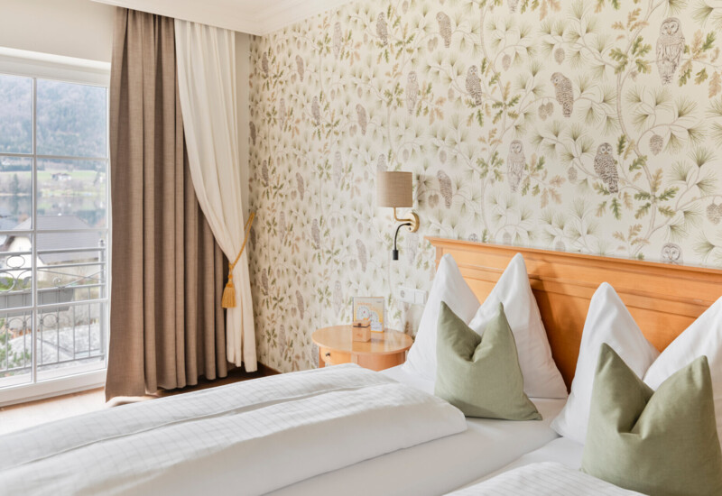 Hotel room with floral and owl wallpaper, a double bed with green pillows, and a scenic view of the lake and mountains through a large window.