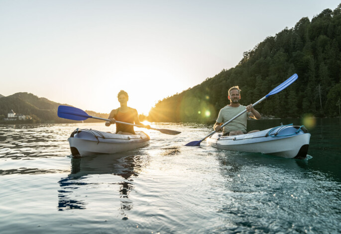 Two people kayaking on lake Fuschl at sunset, surrounded forested hills.