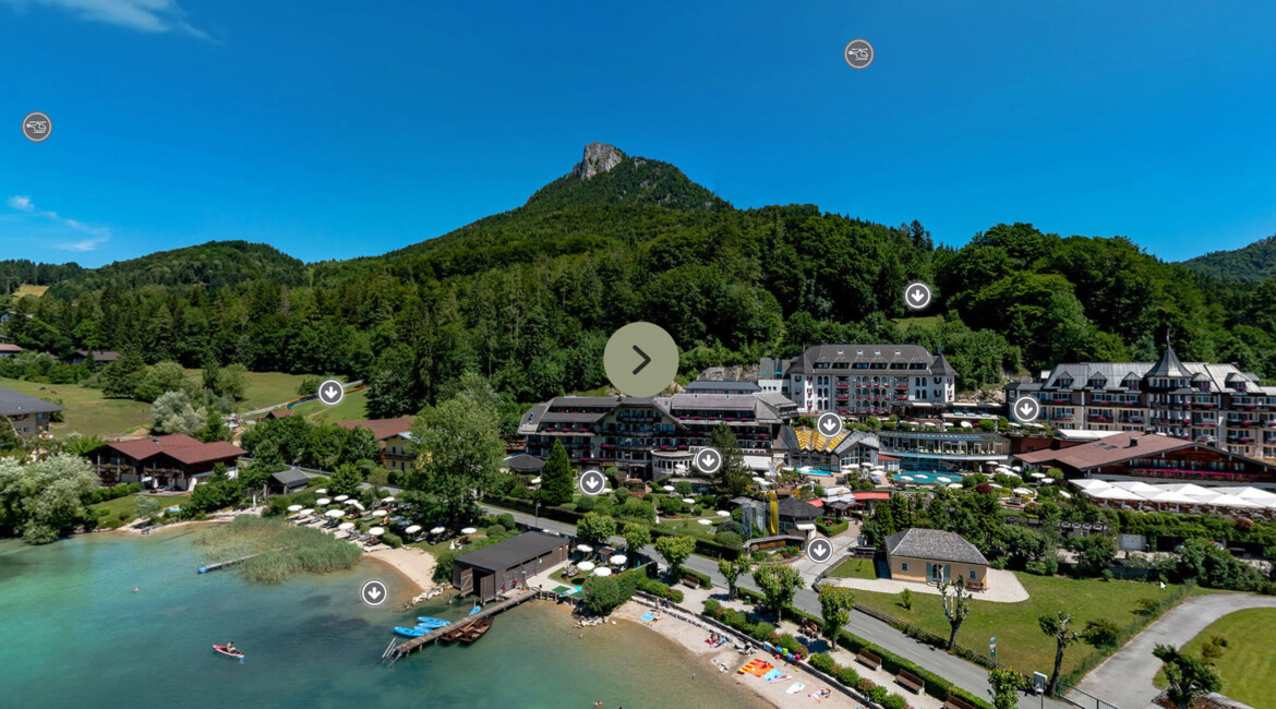 Aerial view of Waldhof Fuschlsee Resort by a lakeside, surrounded by greenery and mountains under a clear blue sky.