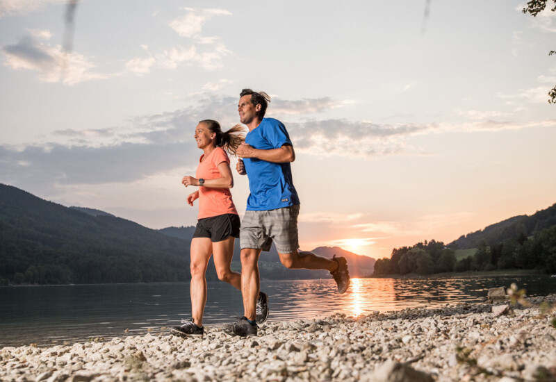 A couple jogging along a lakeside trail at sunset, with scenic mountains in the background.