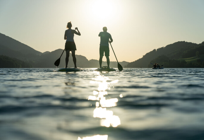 Two people paddleboarding on a tranquil lake at sunset, with hills silhouetted in the background.