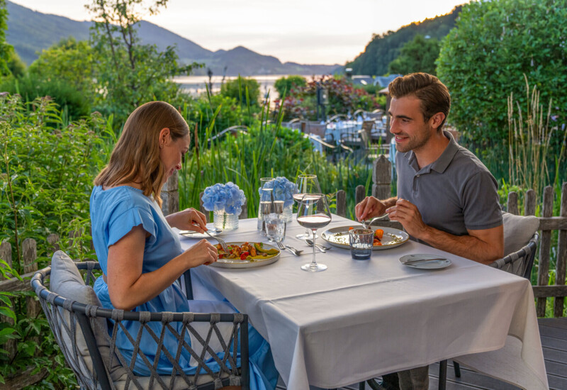 Ein Paar genießt ein Abendessen auf der Terrasse mit Blick auf die malerische Landschaft und den Fuschlsee bei Sonnenuntergang.