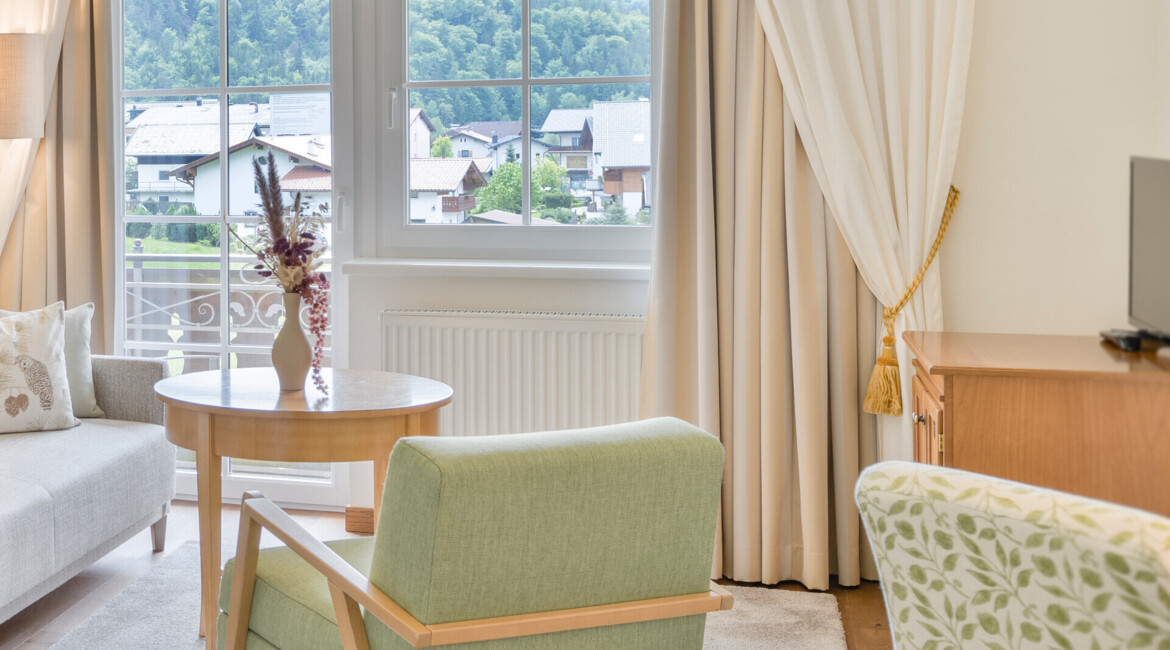 Hotel room with green chair, beige sofa, and a view of lush, scenic mountains through large windows.