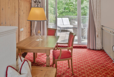 Cozy room with wooden table and red chairs, overlooking a lush balcony with sun loungers through glass doors.