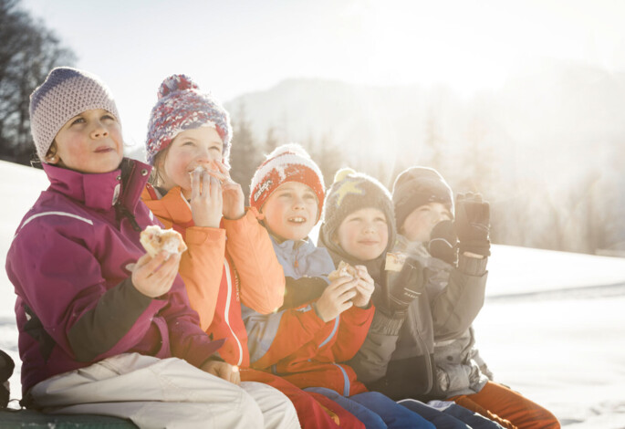 Children wearing winter clothes eating snacks outdoors in snowy weather.