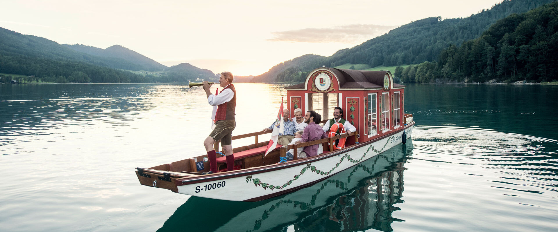 A boat with people in traditional attire sails on a serene lake surrounded by lush, scenic mountains at sunset.