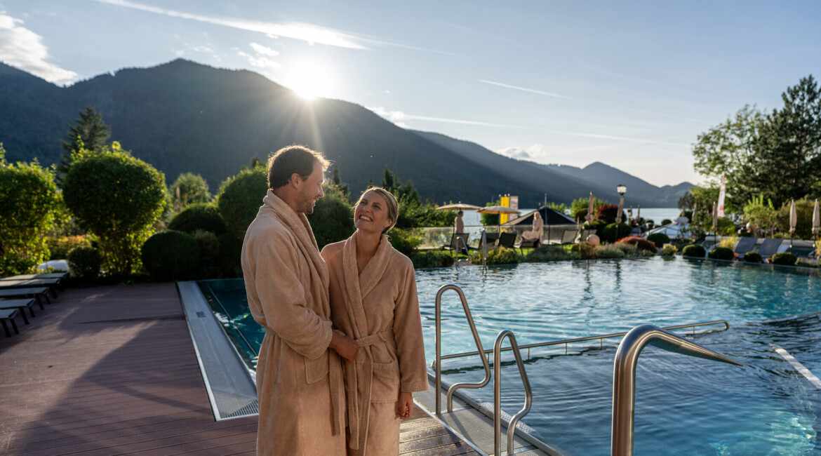 Ein Paar in Bademänteln genießt die Aussicht am Pool vor einer bergigen Landschaft bei Sonnenuntergang.
