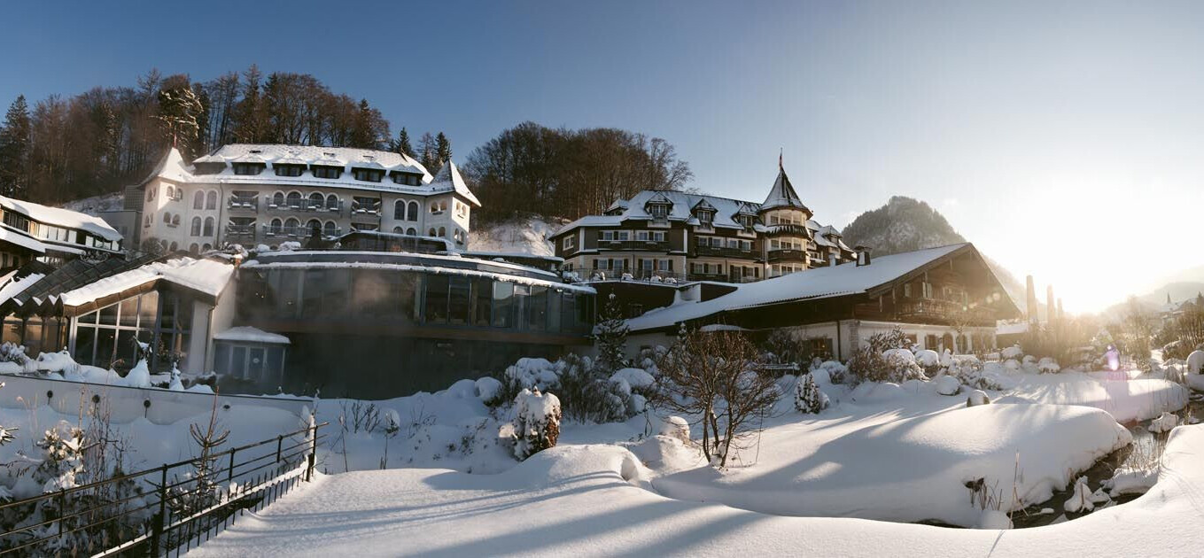 Verschneites Hotelgebäude in malerischer Winterlandschaft bei Sonnenaufgang, umgeben von Bäumen und Bergen.
