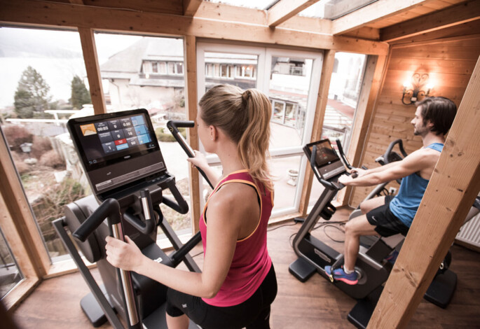 People exercising on machines in a bright, wooden gym with large windows.