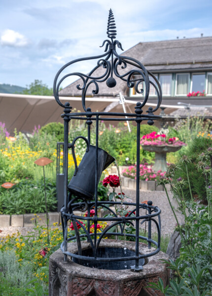 Ein kunstvoller Gartenbrunnen mit schwarzem Metallgestell und Gießkanne im Kräutergarten vom Waldhof Fuschlsee.