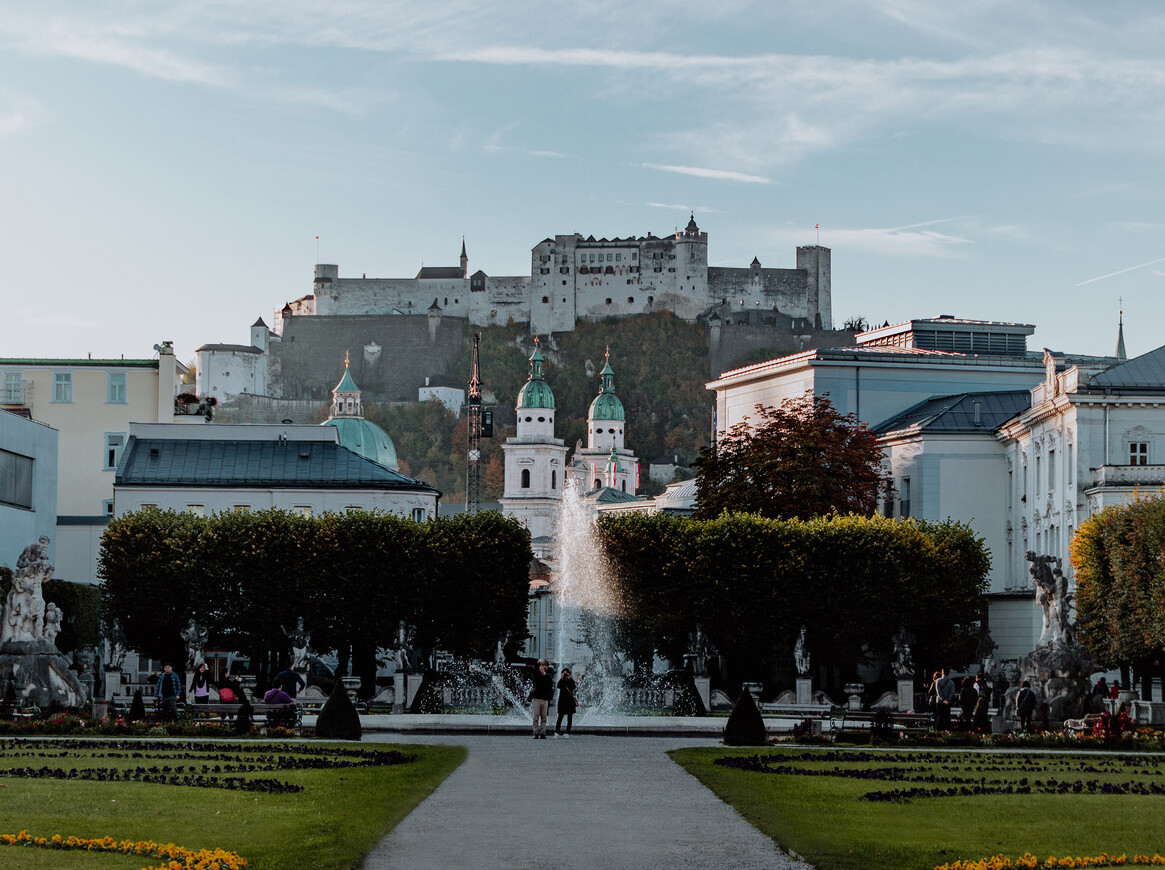 Scenic view of a lush garden with a fountain and a historic fortress in the background, under a clear sky.