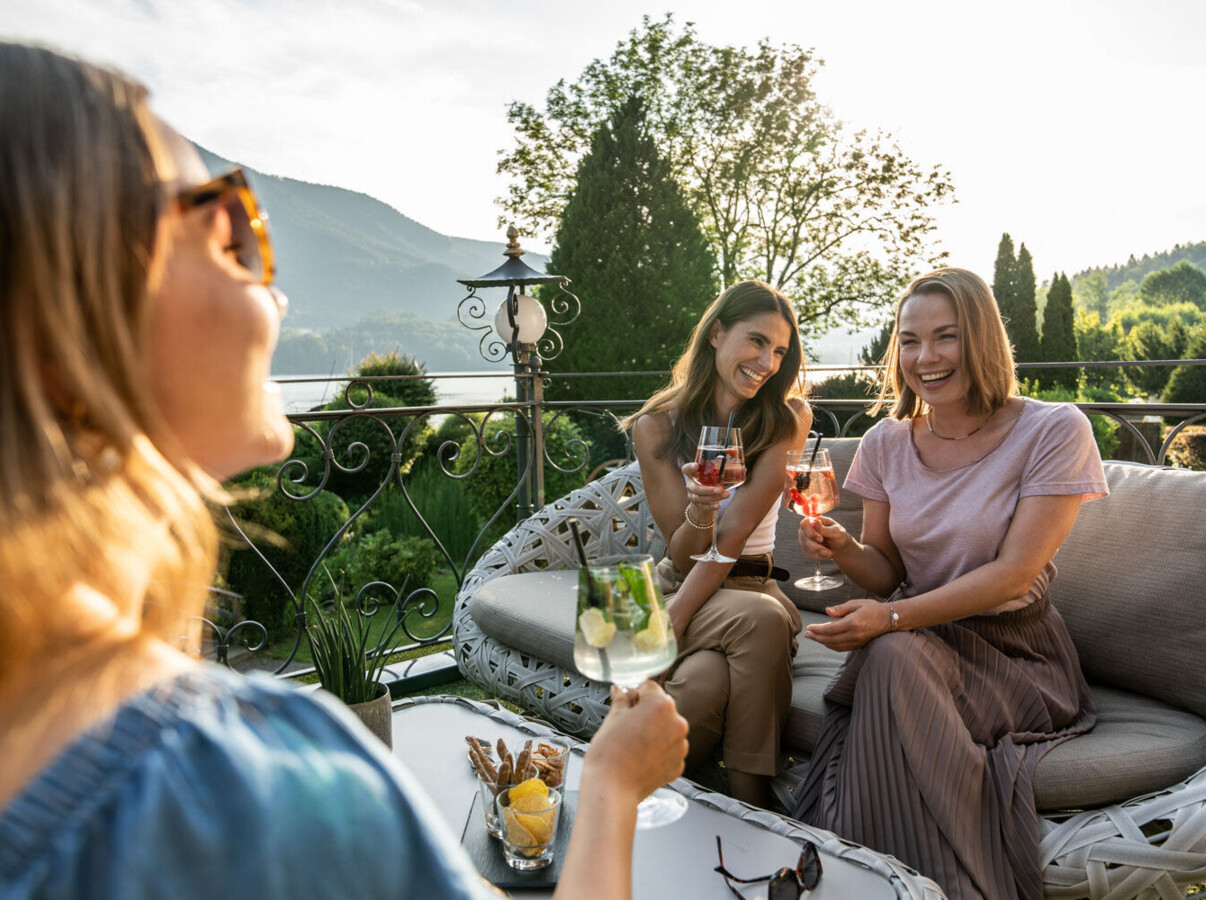 Three women enjoy drinks on a scenic terrace with lush greenery in the background.