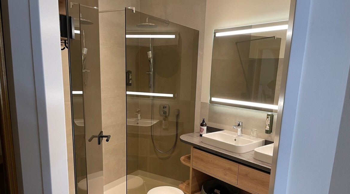 Moderne Badezimmer mit Glasdusche, beleuchtetem Spiegel, Waschbecken und Holzregalen im Hotel Waldhof am Fuschlsee.