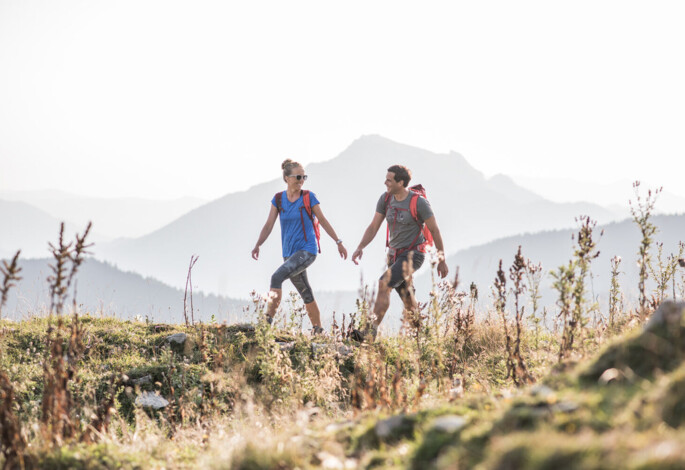 Two hikers with backpacks walking on a grassy trail with mountains in the background.