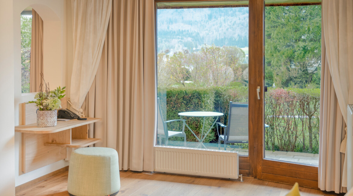 A cozy room with large windows overlooking a lush garden and scenic mountains, featuring a small patio with a table and chairs.