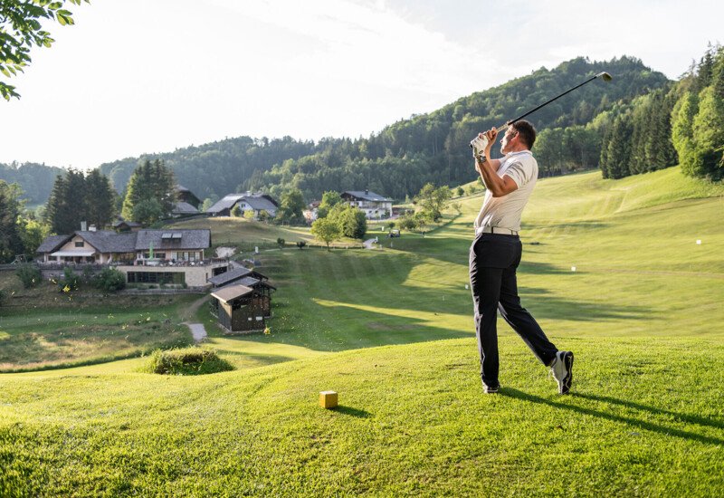 Golfer swings club on lush, scenic course with picturesque buildings and forested hills in the background.