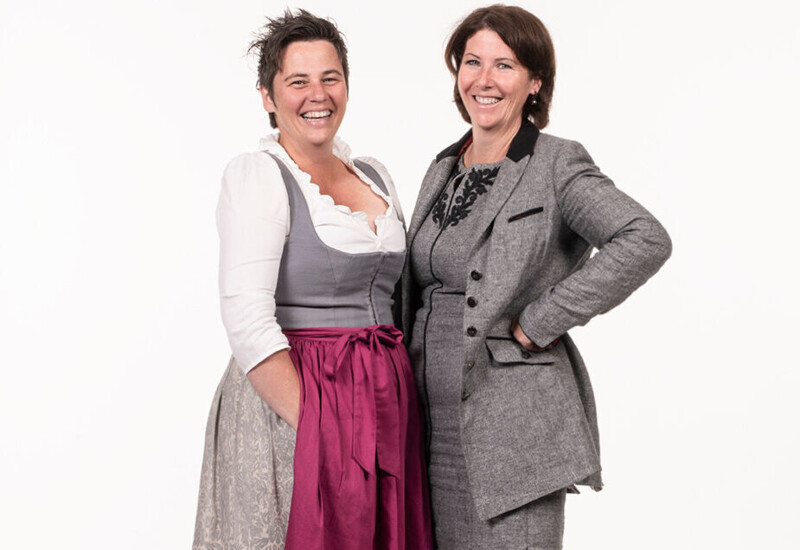 Two women smiling, one in traditional attire with a pink apron, and the other in a gray suit, standing together on a plain background.