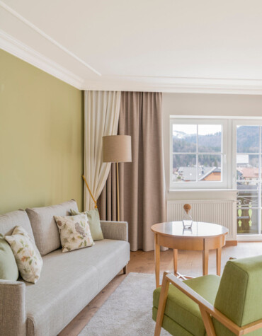 Bright living room with green accents, cozy sofa, armchair, and wooden table by a window offering scenic views.