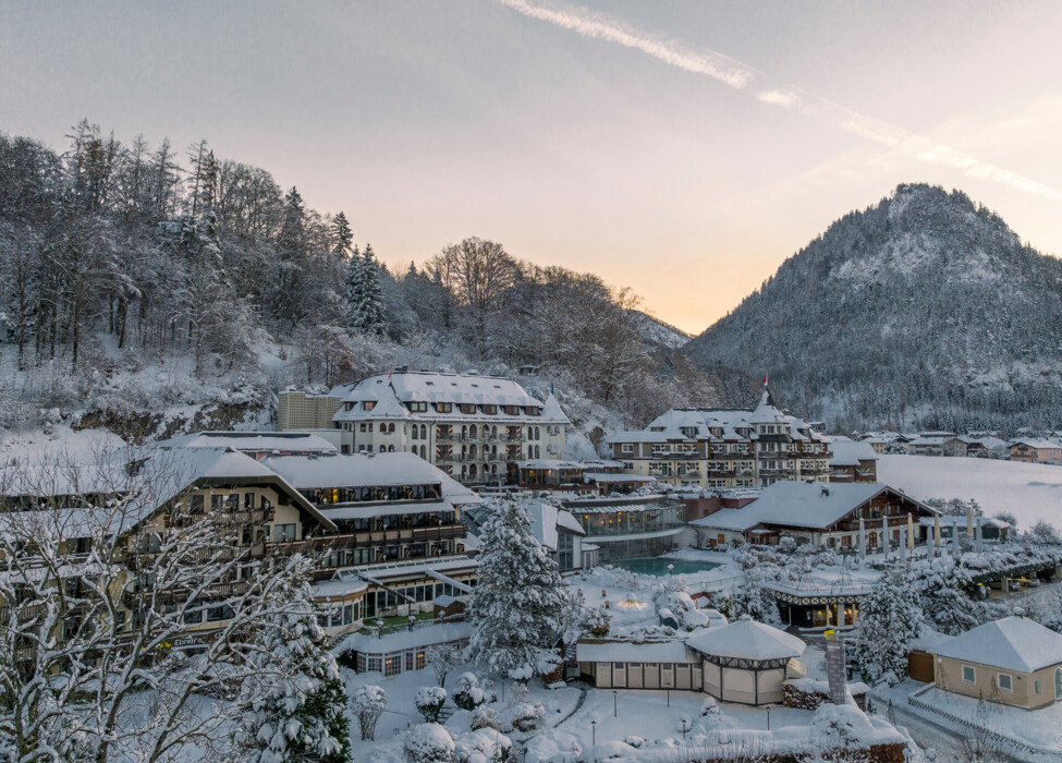 Snow-covered Waldhof Fuschlsee Resort with forested hills and a mountain in the background at sunrise.