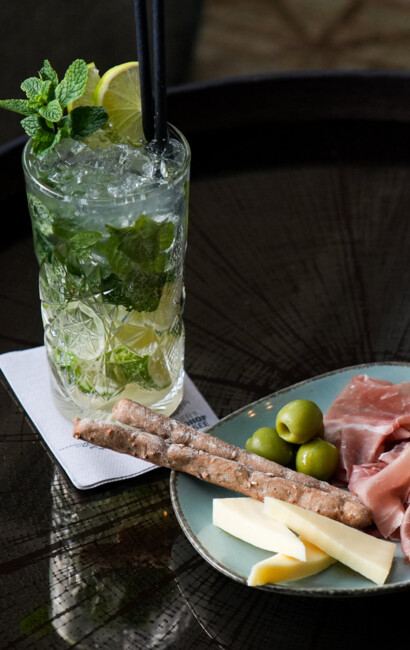 A refreshing drink with mint and lime, alongside a plate of prosciutto, cheese, olives, and breadsticks on a table.