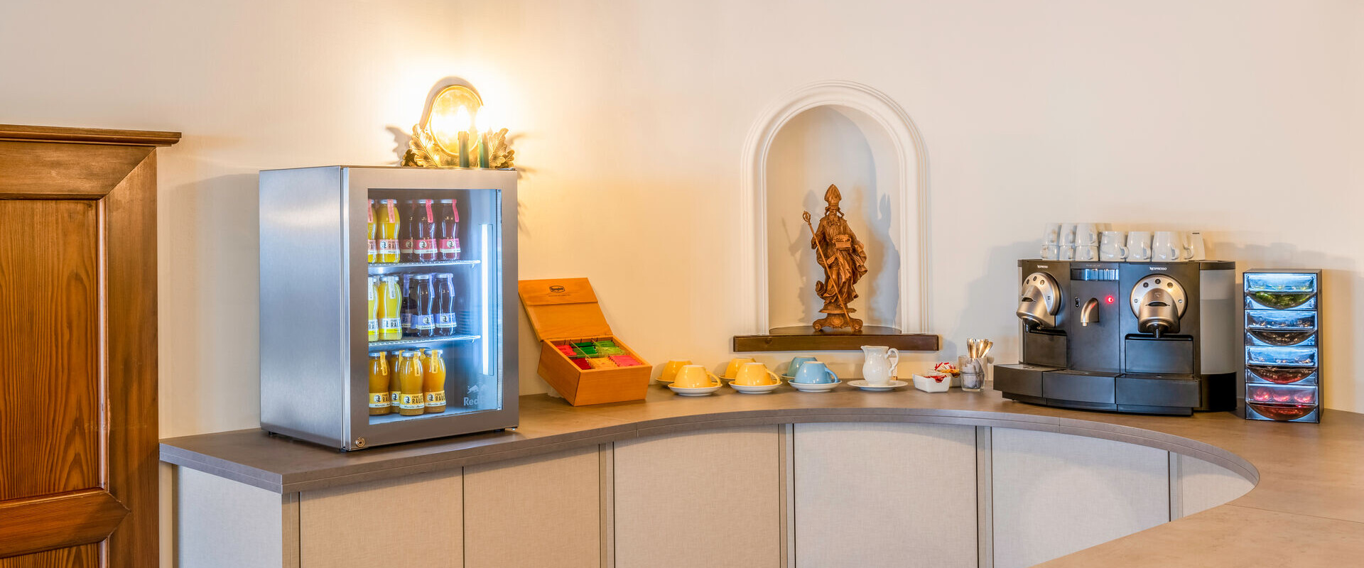 A cozy refreshment corner with a beverage fridge, coffee machine, and cups on a countertop, featuring a decorative statue in a niche.