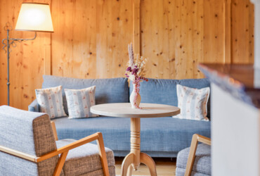 Cozy seating area with wooden walls, featuring a round table, two armchairs, and a blue sofa adorned with patterned cushions.