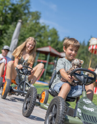 Children enjoying pedal go-karts at Waldhof Fuschlsee Resort, with a carousel in the background.