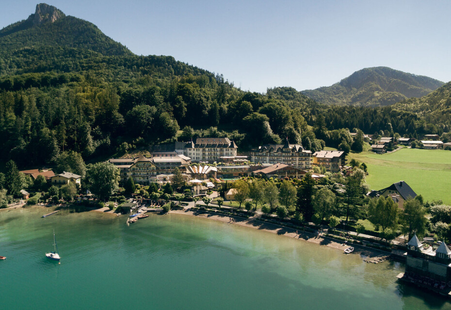 Aerial view of the Waldhof Fuschlsee Resort by a tranquil lake, surrounded by forested hills and mountains.