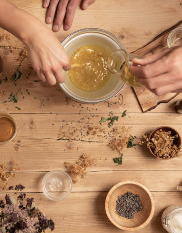 Hands mixing herbs and oils on a wooden table, surrounded by bowls of dried ingredients and bottles, creating a natural remedy.