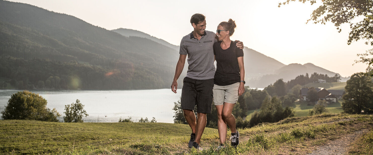 Couple walking on a scenic lakeside path near lake Fuschl with mountains in the background.
