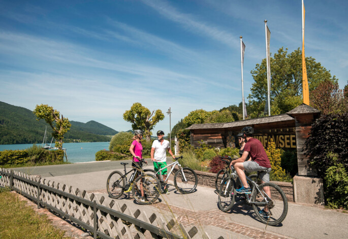 Drei Radfahrer vor der Auffahrt des Waldhof Fuschlsee Resorts, mit dem Fuschlsee im Hintergrund. 