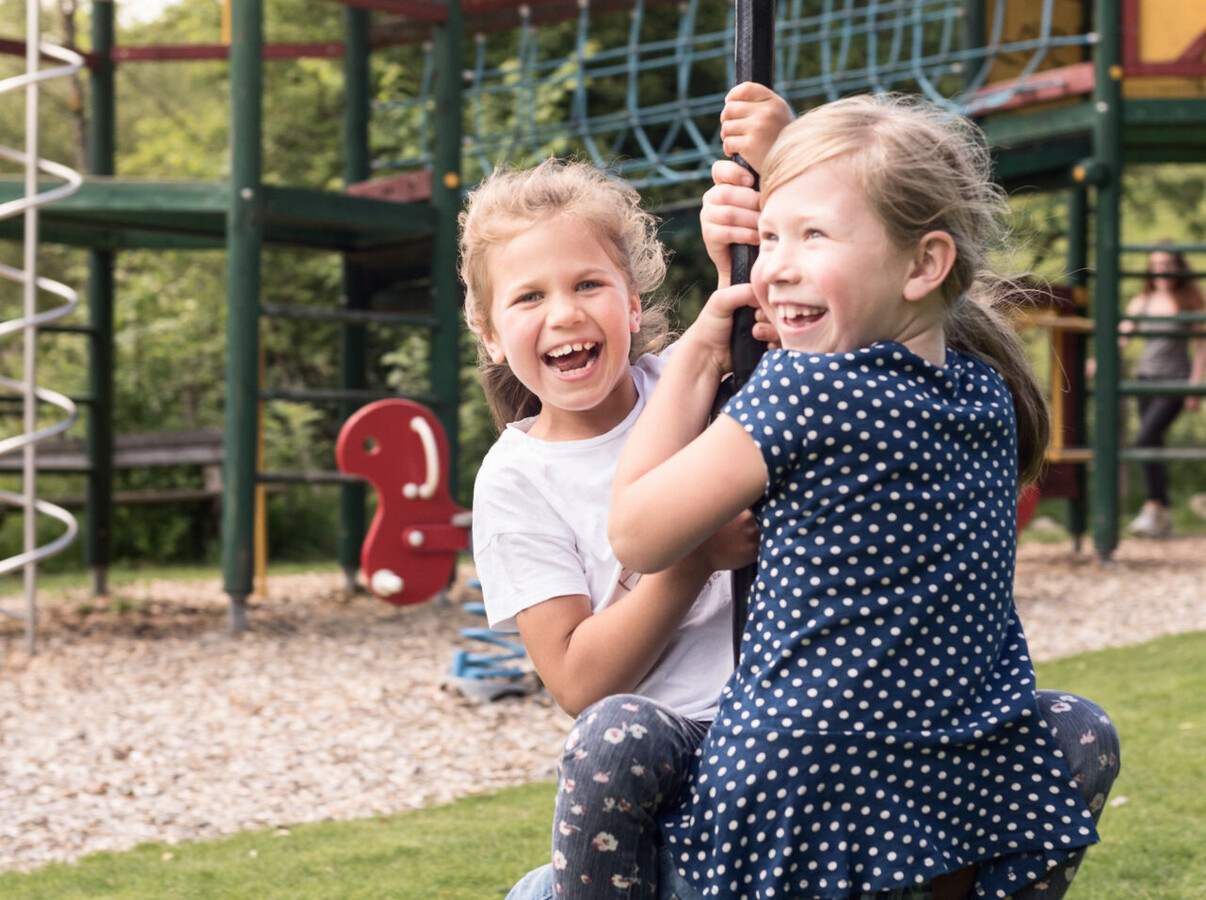 Two children laughing on a swing in a playground surrounded by lush greenery.