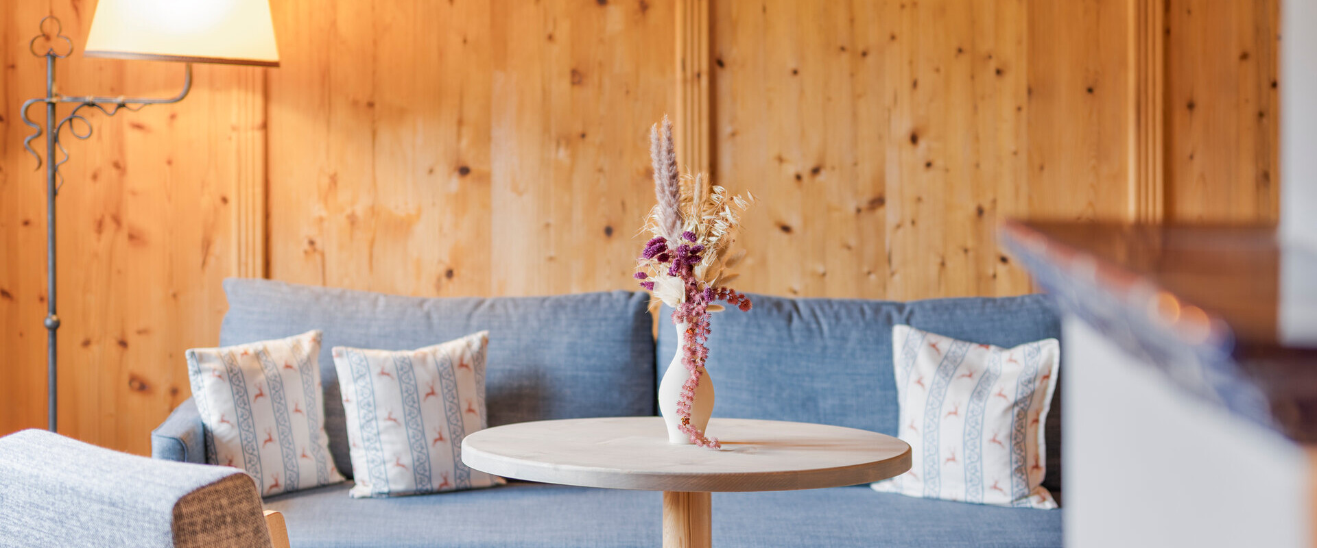 Cozy seating area with wooden walls, featuring a round table, two armchairs, and a blue sofa adorned with patterned cushions.