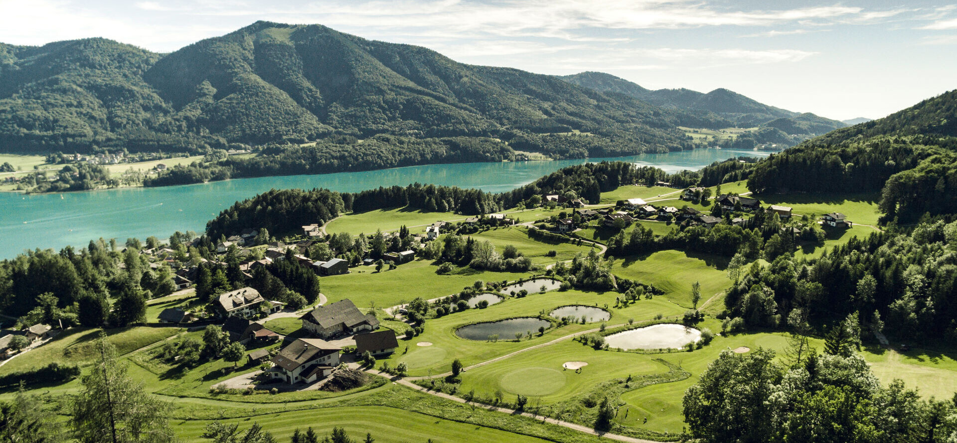 Scenic aerial view of a lush golf course surrounded by mountains and a lake.