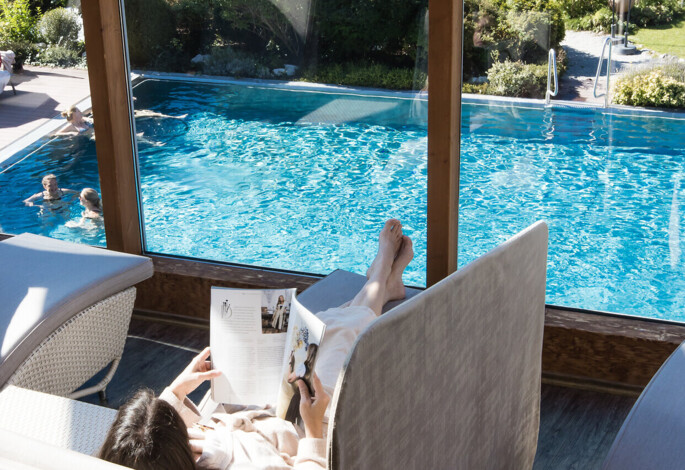 A person relaxes on a lounger with a magazine, overlooking an outdoor pool and garden at Waldhof Fuschlsee Resort.