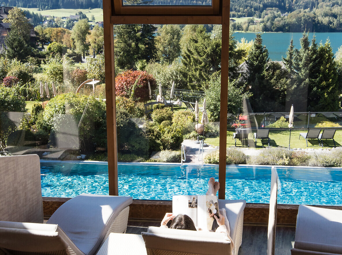 Eine Person entspannt auf einer Liege am Pool mit Blick auf Berge und einen See durch eine Glasfront.